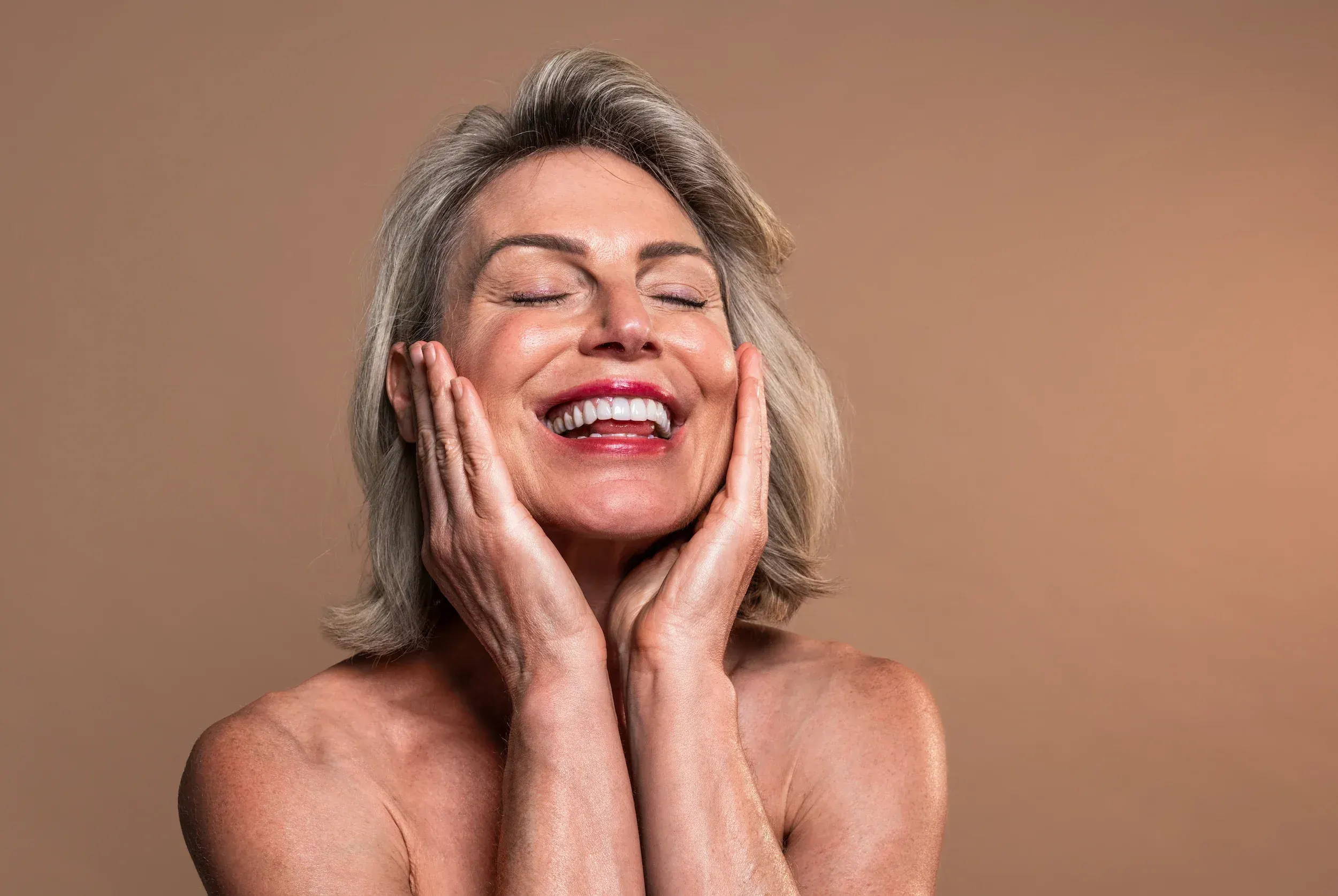 Woman with silver-streaked hair smiling with eyes closed touching her face - embracing healthy radiant skin at every age